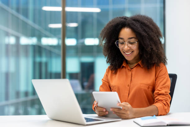 istock Young professional woman smiling while working on digital tablet and laptop in modern office setting 2187317696