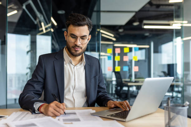 istock Young professional working in modern office focused on paperwork and using laptop. Businessman wearing glasses seated at desk surrounded by documents, colorful sticky notes visible on glass wall. 2187323889