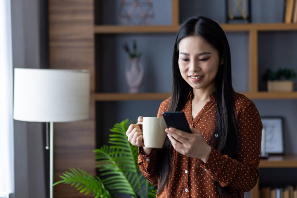 istock Asian woman in cozy home setting enjoying coffee while texting on smartphone, wearing brown polka dot blouse, surrounded by modern decor, showcasing relaxed lifestyle and modern communication. 2187327552