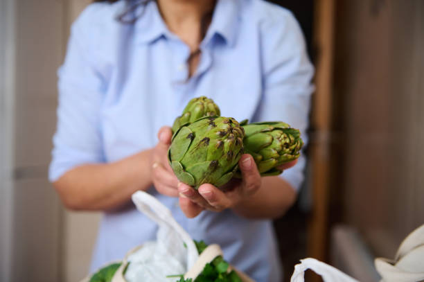A person presents fresh artichokes in their hands, symbolizing healthy eating and culinary freshness. The image captures a homey kitchen atmosphere, emphasizing organic and natural food choices.