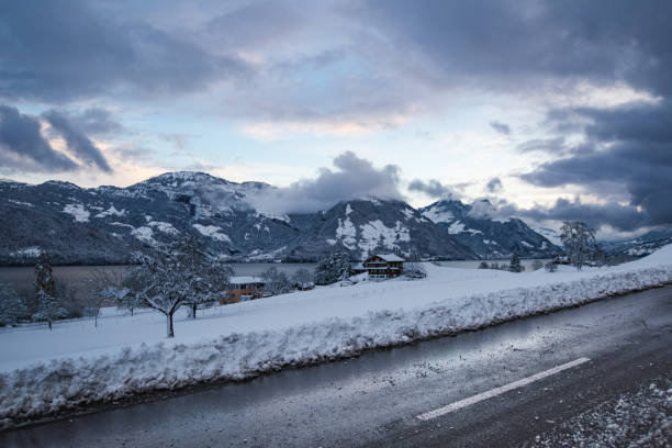 A picturesque winter landscape featuring a freshly plowed road bordered by thick snowbanks. In the distance, serene mountains and a tranquil lake are framed by dramatic clouds and snow-laden trees.