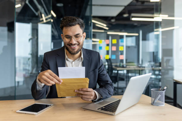 Business professional opening envelope while sitting at desk in modern office with laptop, tablet, and paperwork, looking pleased with correspondence received during workday