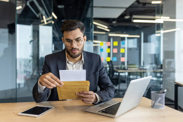 istock Serious man in a modern office holding a postal envelope, reading an important message. He is focused and appears thoughtful, surrounded by technology and office materials. 2187338920