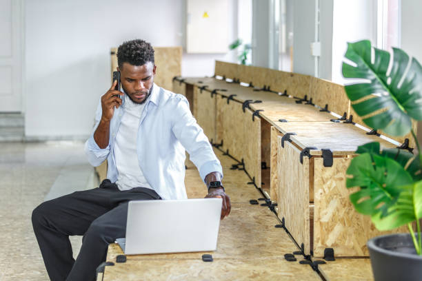 A young man is focused on a business call while sitting at a wooden table and using his laptop in a contemporary office space. Green plants add a touch of nature to the environment.