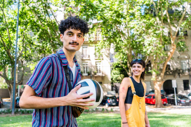 Group of four friends, diverse in ethnicity including an African-American man, a Hispanic woman, a non-binary White individual, and an Asian man, engaging in a playful game of volleyball on sunny day in a park. They are seated on grass, surrounded by trees, and appear to be having fun with a volley ball that a man in blue shirt is trying to catch.
