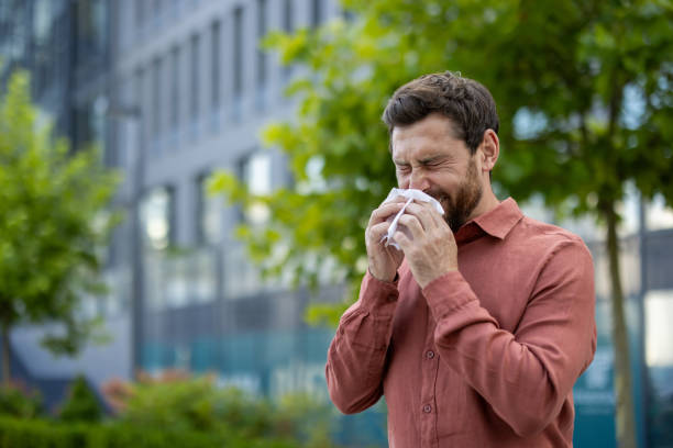 Mature man sneezing in urban environment captured in clear focus, surrounded by greenery and modern architecture, evokes feelings of discomfort and seasonal allergies on city streets