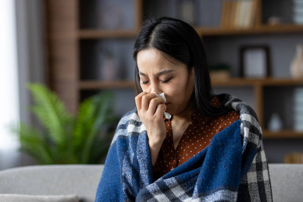 istock Asian woman at home experiencing cold symptoms, covered with warm blanket, holding tissue, showing signs of illness, feeling unwell, seeking comfort and warmth indoors 2187349934