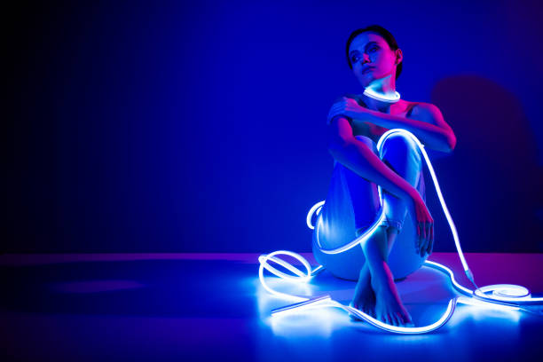 istock Young woman in neon lights sitting against dark blue background in studio 2187353236