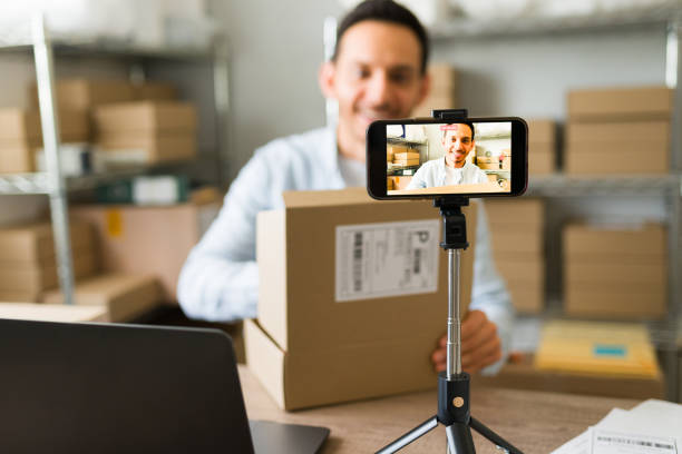 istock Smiling business owner with a package, creating a promotional video for his online store using a smartphone 2187360709
