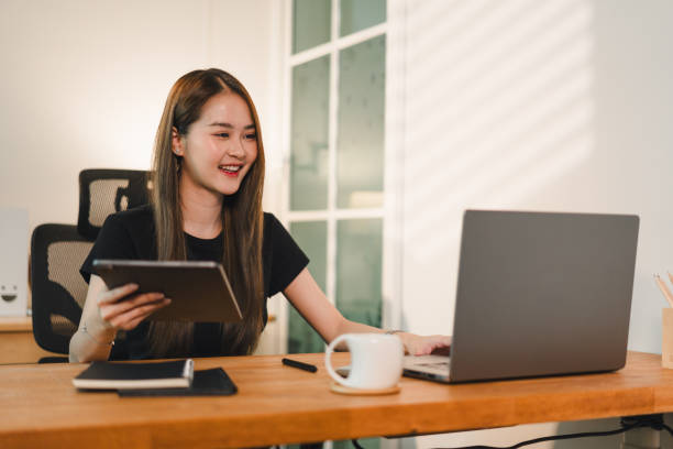 istock young woman is working on laptop while holding tablet, smiling and engaged in her task. modern workspace features wooden desk and cup of coffee, creating productive atmosphere 2187363560