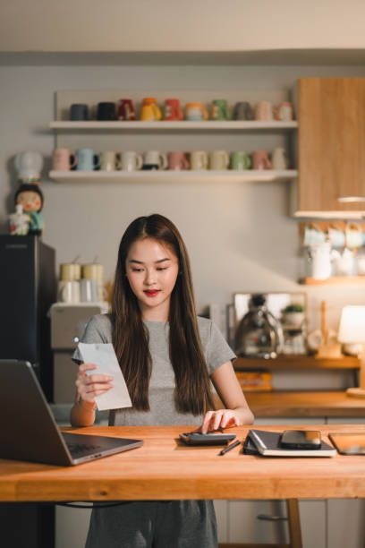 istock young woman is sitting at wooden table in cozy kitchen, reviewing document while using laptop. background features colorful mugs on shelf, creating warm and inviting atmosphere 2187363612