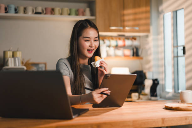 istock Enjoying snack while working on tablet, young woman sits at wooden kitchen table. bright, sunlit space features laptop and various kitchen items, creating cozy and productive atmosphere 2187363632