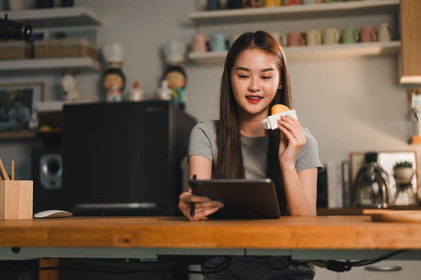 istock Enjoying snack while using tablet, young woman sits at wooden table in cozy kitchen. scene captures moment of relaxation and engagement with technology 2187363651