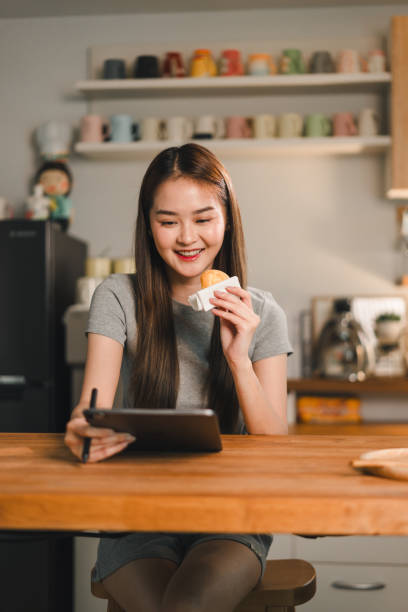 istock young woman enjoys snack while using tablet in cozy kitchen. warm atmosphere and her cheerful expression create delightful scene of relaxation and productivity 2187363656