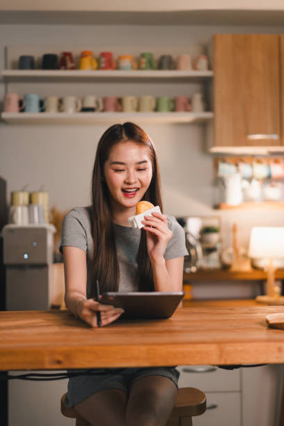 Enjoying snack while using tablet, young woman smiles in cozy kitchen filled with colorful mugs. warm ambiance creates cheerful and relaxed atmosphere