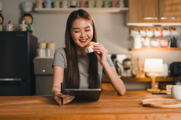 istock Enjoying snack while using tablet, young woman smiles in cozy kitchen setting. warm ambiance and her relaxed demeanor create delightful moment of leisure 2187363659
