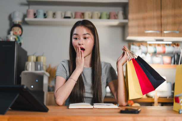 istock Surprised woman holding shopping bags while sitting at table with notebook and tablet. She expresses excitement and curiosity about her purchases 2187364350