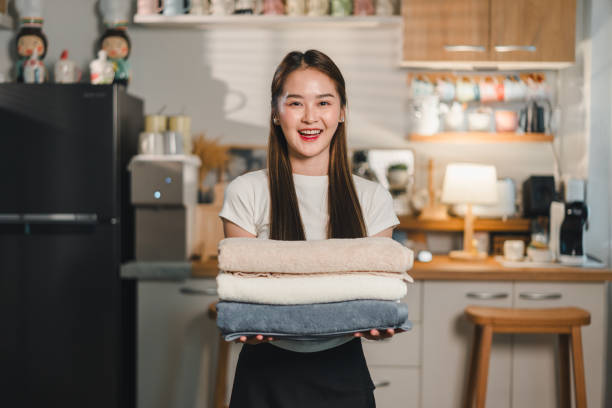 young woman smiles while holding stack of neatly folded towels in cozy kitchen setting. warm ambiance and her cheerful expression create welcoming atmosphere