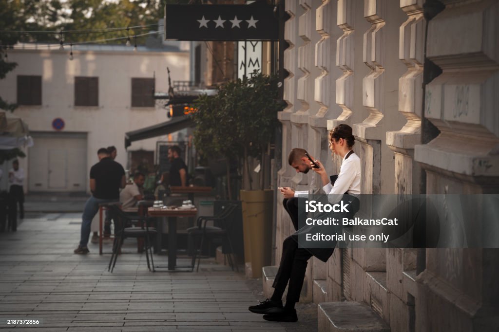 Deux serveurs, un homme et une femme, assis à l’extérieur pendant une pause et vérifiant leurs smartphones, illustrant l’utilisation de la technologie moderne par les travailleurs de l’hôtellerie pendant les temps morts. - Photo de Assis libre de droits Deux serveurs, un homme et une femme, assis à l’extérieur pendant une pause et vérifiant leurs smartphones, illustrant l’utilisation de la technologie moderne par les travailleurs de l’hôtellerie pendant les temps morts. - Photo de Assis libre de droits