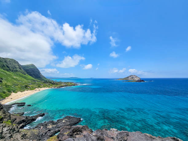 A wide view of the sea and mountains, seen from a high vantage point, with a clear blue sky and scattered clouds enhancing the scenery.