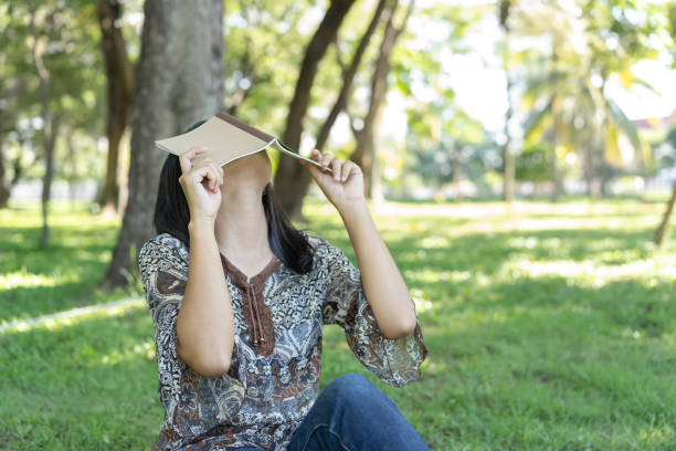 A young woman covers her face with a book while sitting on the grass with her head tilted back, resting from exhaustion.