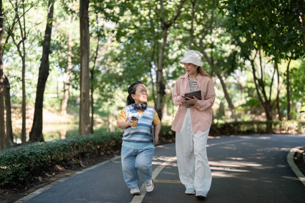 A young person with dwarfism and a friend enjoying a conversation while walking in a green, sunny park.