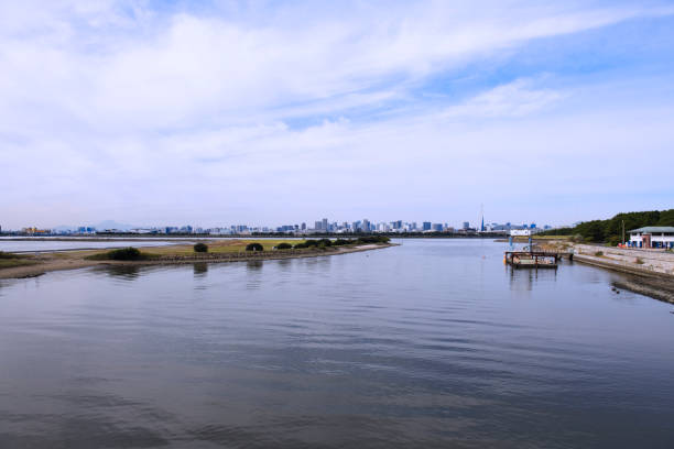 A passenger boat travels across the Chao Phraya River with a cityscape of Bangkok in the background under an overcast sky. The urban skyline includes a modern high-rise under construction, contrasted with traditional Thai architecture.