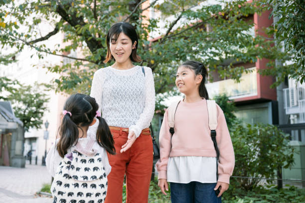 Japanese mother smiles at her two young daughters while walking down a sidewalk in tokyo, japan, enjoying a cheerful conversation together after school in the afternoon