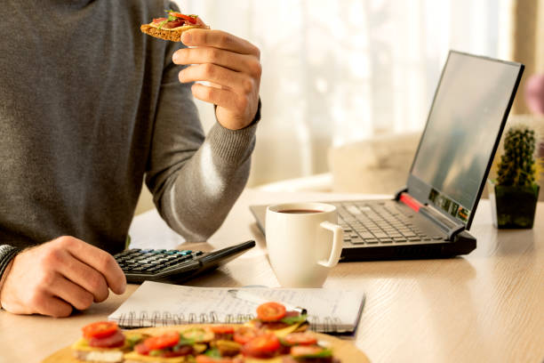 Man snacking on sandwich and coffee while working at table in office on laptop