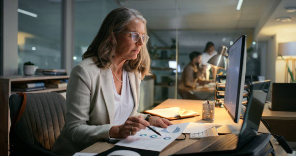 istock Computer, documents and night with business woman in office for budget or financial statistics. Accounting, bookkeeping or information and mature employee in workplace with finance charts or graphs 2187408428