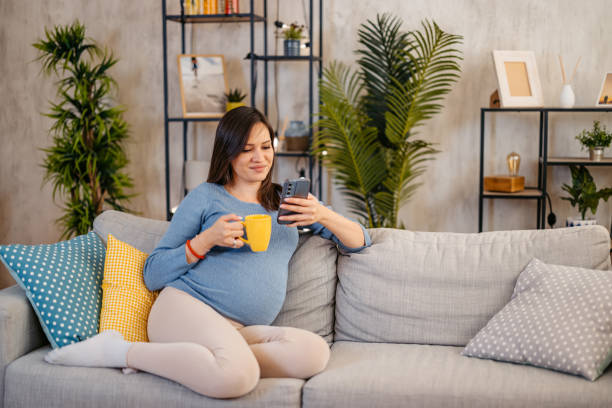 istock Young Pregnant Woman Sitting On The Sofa And Using Phone While Drinking Coffee 2187436933