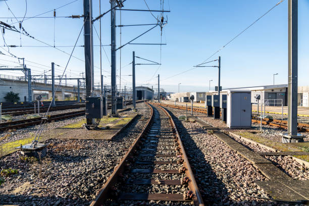 Rusty railway tracks are converging at a train depot, featuring electrical infrastructure and control boxes under a clear blue sky