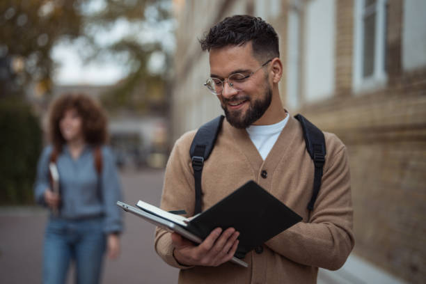 istock Smiling student with glasses and a backpack, holding an open notebook outdoors 2187460698