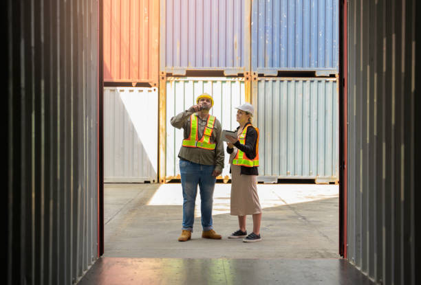 istock Two workers wearing safety vests and hard hats while standing in shipping container yard. Male holding walkie talkie and female using digital tablet for cargo loading information 2187464747