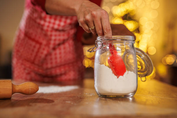 A person in a red apron scoops flour from a glass jar with a red spatula, evoking warmth and holiday baking. Soft, glowing lights create a festive, cozy kitchen atmosphere.
