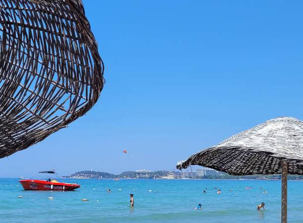 Aydın, Turkey - July 18, 2024: People enjoying near the sea at beach on summer holiday. Some of them swimming and others are parasailing on Aegean sea, you see town of Kuşadası in Turkey on a sunny summer day, View Of Kusadasi Skyline At Aegean Coast, Turkey.