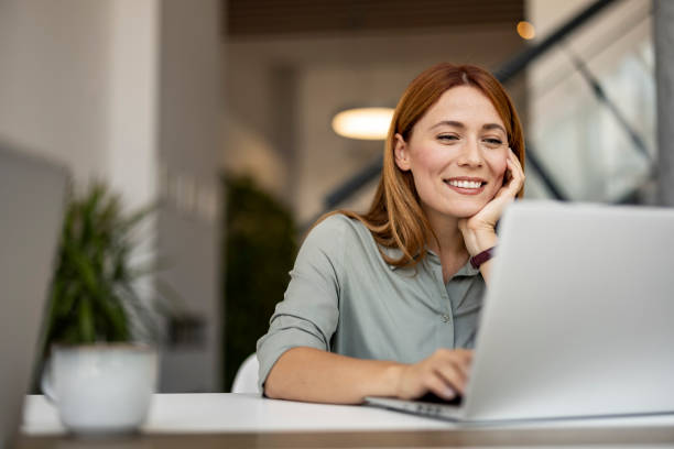 Smiling Woman Working Remotely on Laptop in Modern Workspace A smiling woman sits at a desk, working on her laptop in a contemporary, well-lit workspace. She appears engaged and content, suggesting a positive remote working experience. using computer stock pictures, royalty-free photos & images