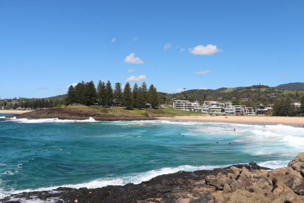 istock Coastal view with sandy beach, surfers, and lush greenery under a clear blue sky in a scenic seaside town 2187527506