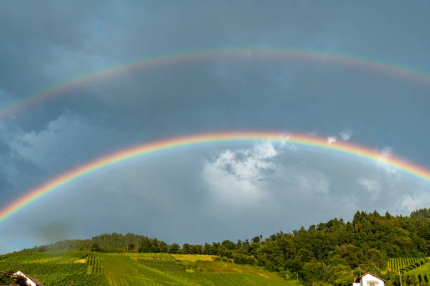 A rainbow is seen in the sky above a hillside. The sky is cloudy and the sun is not visible