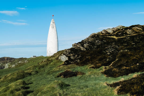 istock A slender, white Beacon stands prominently against the blue sky. It is situated on a hill composed of dark soil, grass, and exposed rocks. 2187554512