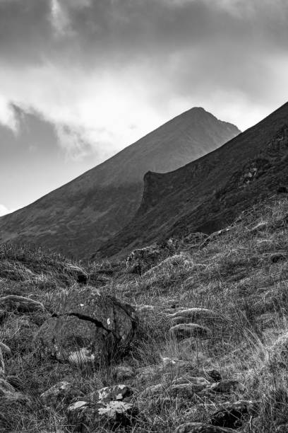 istock A steep, dark-grey mountain dominates the scene under a partly cloudy sky. The foreground features patchy grass and moss covering numerous rocks. 2187554555
