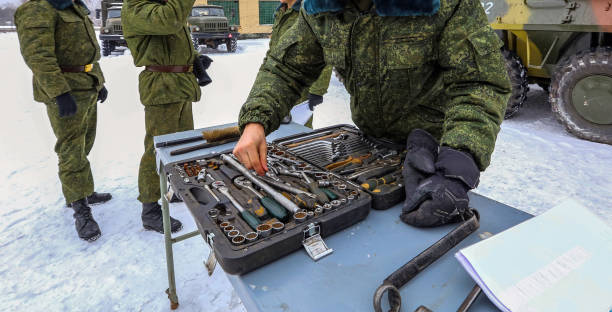 Military personnel inspect tools on a snowy field during a training exercise in winter with vehicles in the background