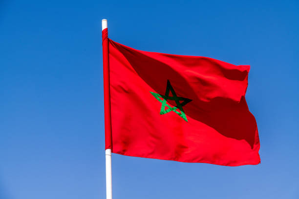 Agadir, Souss-Massa - Morocco - 08-03-2024: A Moroccan flag with a green five-pointed star waves on a white pole against a clear blue sky, appearing dynamic in the wind.