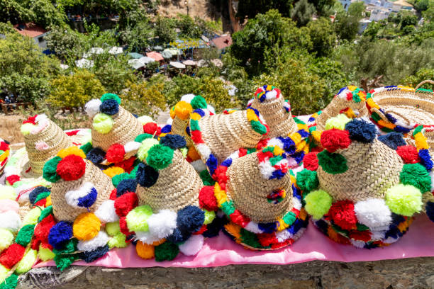 Straw hats with colorful pompons in red, blue, green, yellow, and white on pink cloth, surrounded by greenery. Traditional Moroccan craftsmanship.