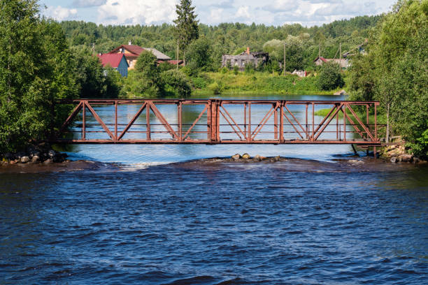 Interesting view of old iron bridge over rapids on river, complex engineering structure against water and green trees, roofs of village houses in the distance, sky and clouds, rivers and lakes, rural landscape, travel and tourism, summer.