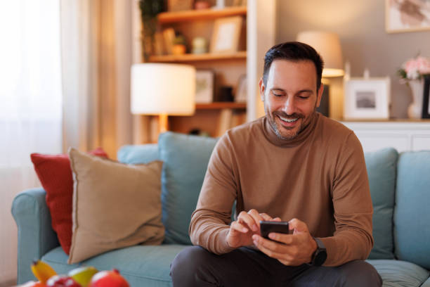 Portrait of handsome man smiling and scrolling apps over smart phone while sitting on sofa at home