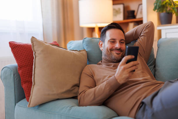 istock Handsome man smiling and messaging over smart phone while resting on sofa in living room at home 2187568819