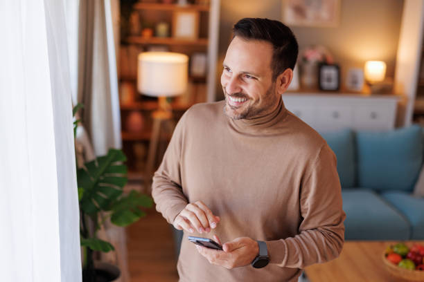 istock Portrait of cheerful handsome man looking through window and using phone while standing in home office 2187568821