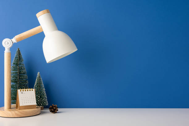 istock A side view cozy winter-themed minimalist desk setup featuring a modern lamp, miniature Christmas trees, a calendar, and a small pinecone against a blue background 2187575593