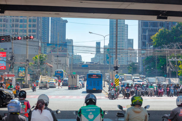 Crowded queued traffic jam and Bangkok cityscape seen from juntion at Rama 9 Road. In foreground thai people on motorcycles are waiting  at junction.  Cars are crossing the junction from left to right. In background is queued traffic jam on Thanon Asok towards Sukhumvit Road. In far background cars and buses are passing over elevated road abive main street Thanon Asok